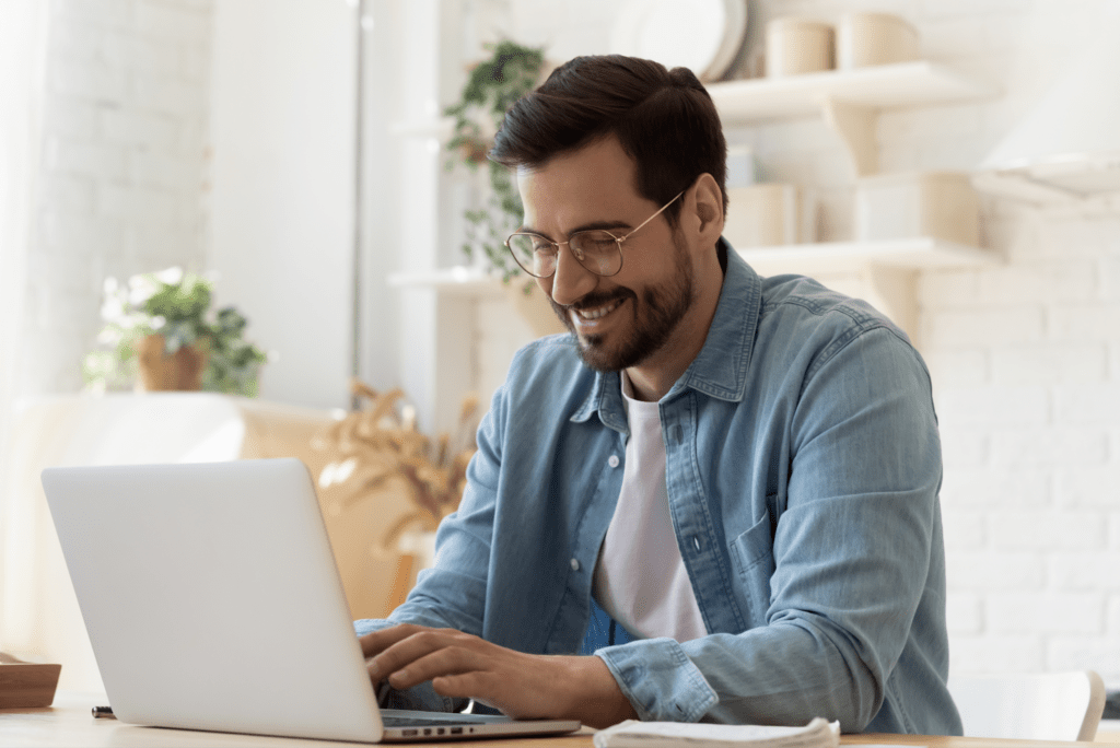 man with glasses smiling whilst working on a laptop